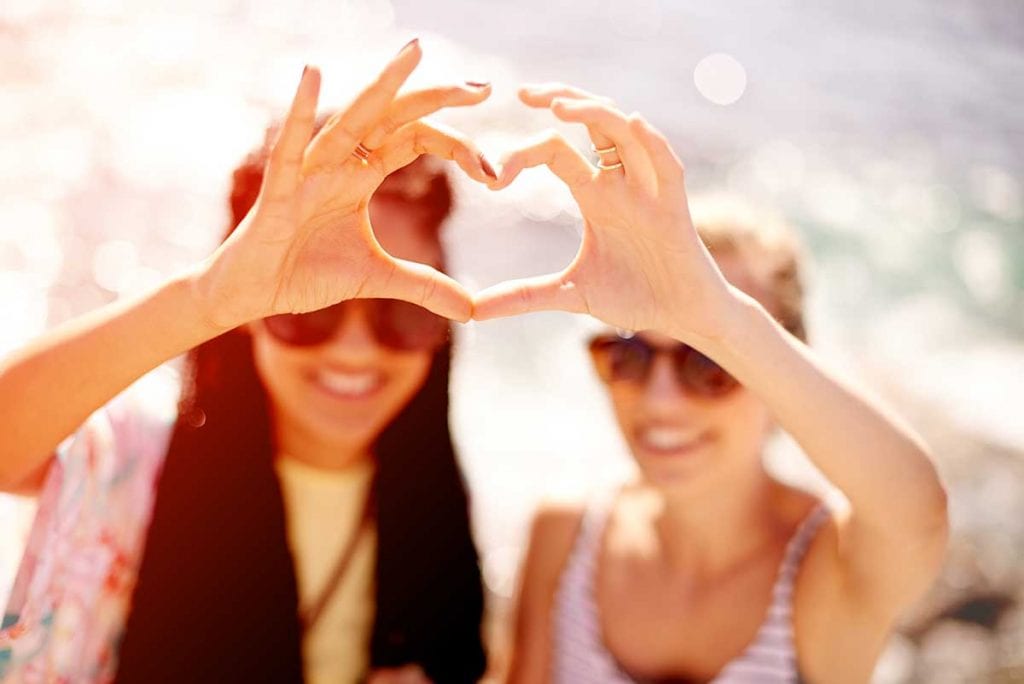 two young female adults outside wearing sunglasses with their hands meeting into a shap of a heart