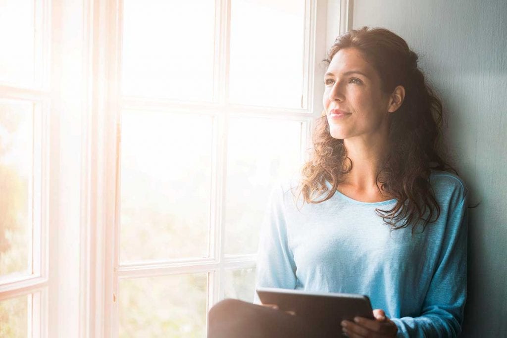 woman holding device sitting on window sill looking out of window