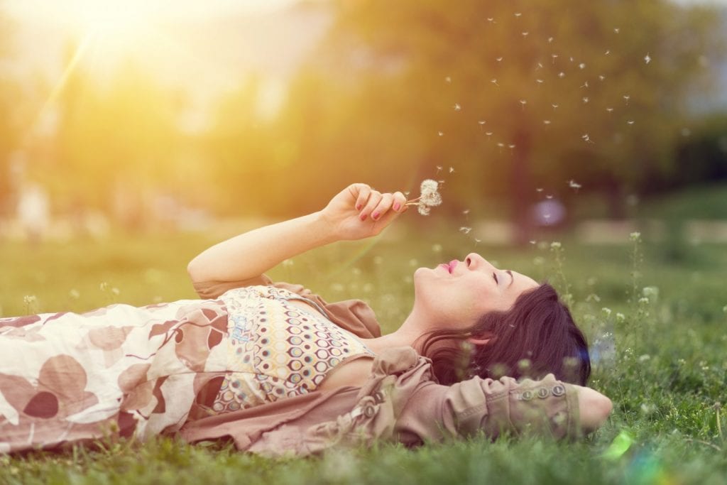 relaxed woman lying in the grass blowing on a bunch of dandelions with sunlight shining