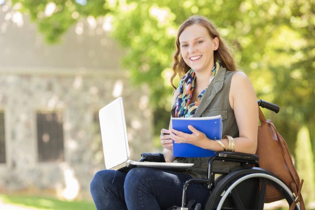 Young female adult sitting in wheelchair with a laptop on her lap and a notebook in her hand