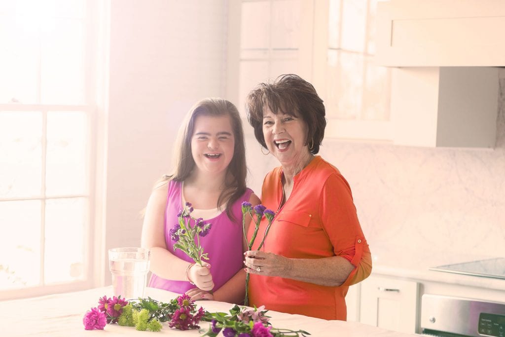 Older happy lady standing next to teenage girl with down syndrome doing a flower arrangement together in a kitchen