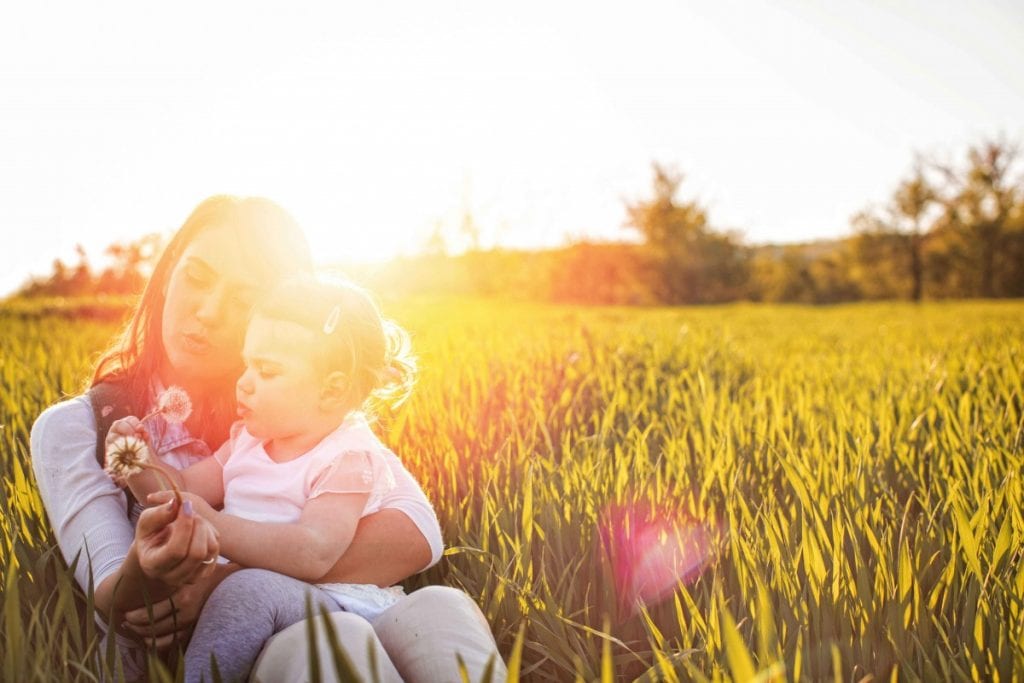 mother sitting in field of grass with young daughter blowing on dandelions